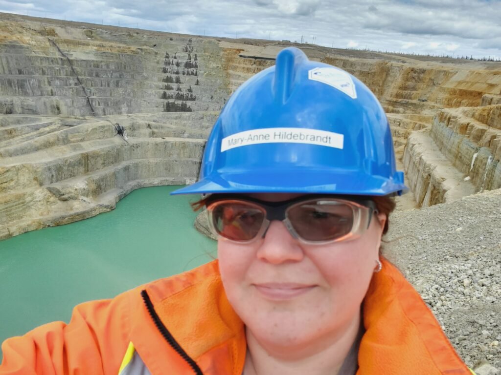 Mary-Anne is holding the camera in selfie-mode and smiling. Behind her there is a large mine with a turquoise lake at the bottom of the cliffs.