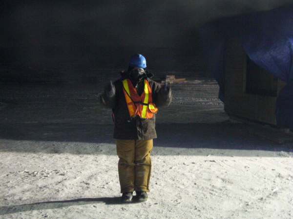 Mary-Anne is giving two thumbs up to the camera. She is wearing protective gear including a helmet and vest. She is lit by a bright light coming from the side and the background is in shadow.