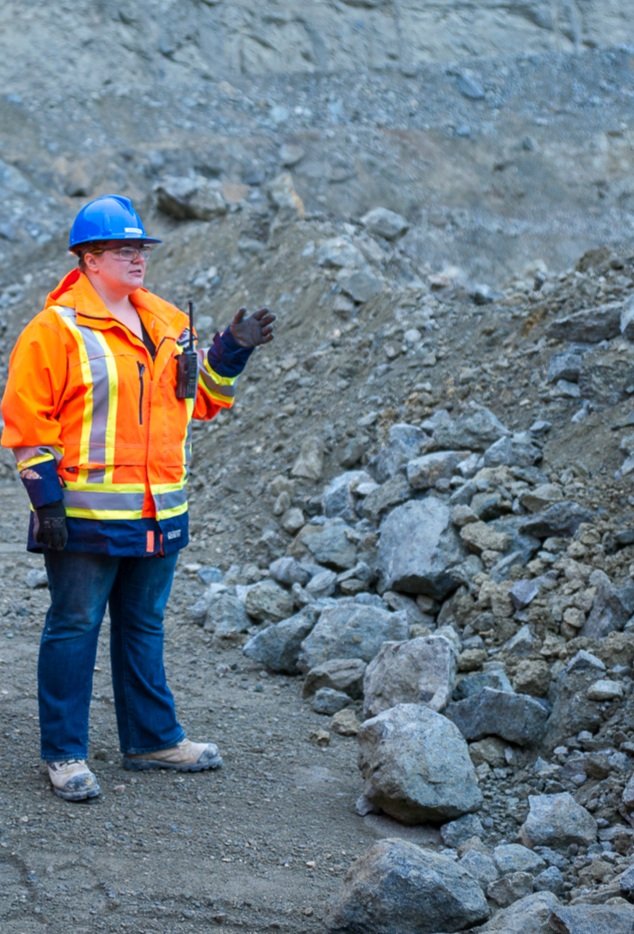 A woman in a reflective orange jacket and blue safety helmet is facing a pile of boulders and gesturing with her hand to the rocks. 