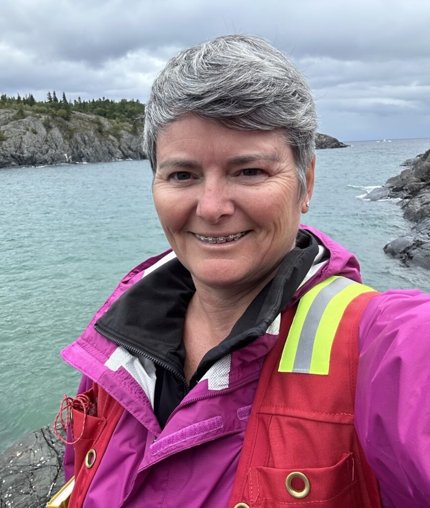 A woman is smiling at the camera wearing a pink jacket and red high visibility vest. Behind her is a body of water with cliffs and trees on either side.