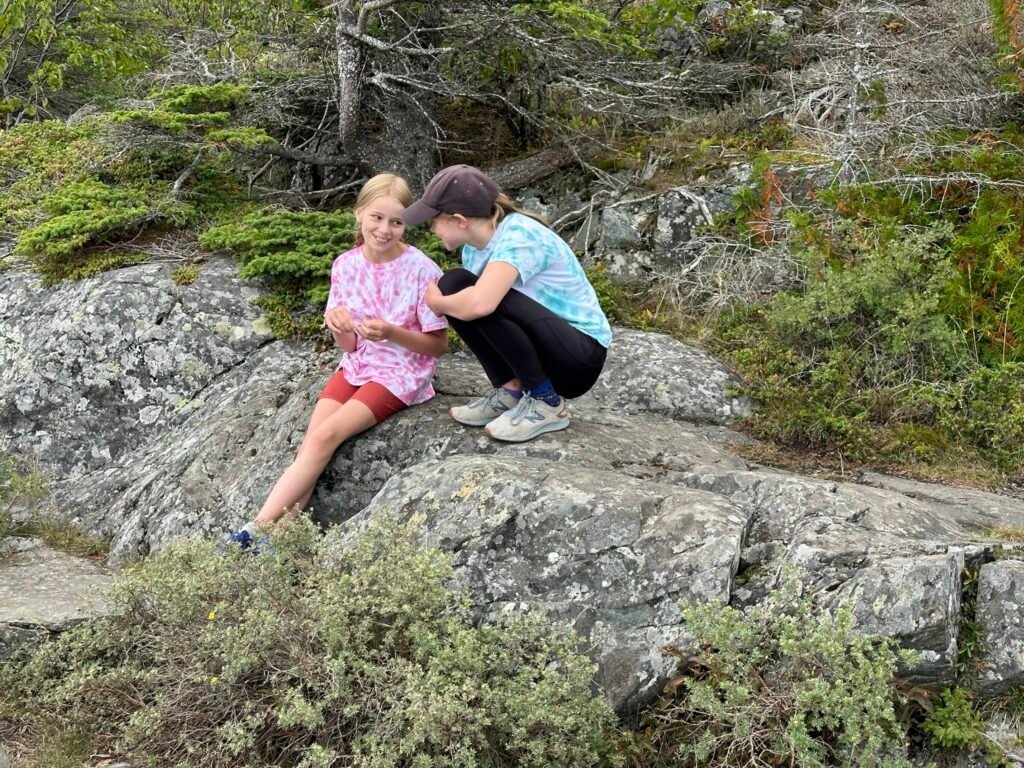Two pre-teen girls sit on a rock outcrop surrounded by trees and bushes. They appear to be talking to one another.