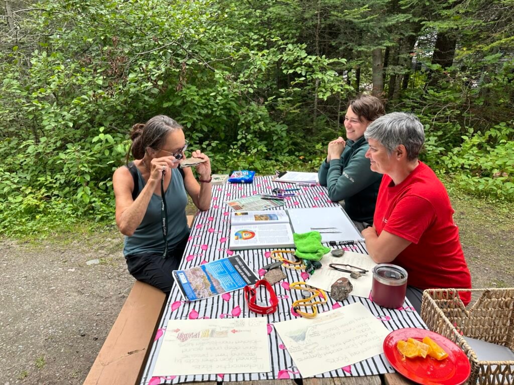 Three adults sit around a picnic table. On the table sit rocks, fossils, information sheets and hand lenses. One of the people is holding a rock to their eye and looking at it through a hand lens (magnifying glass).