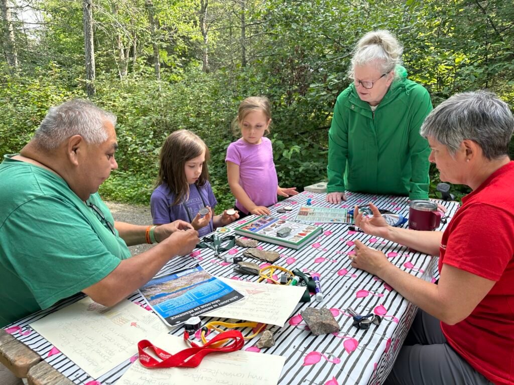 Three adults and two kids sit at a picnic table. On the table are rocks, fossils, information sheets, and hand lenses.