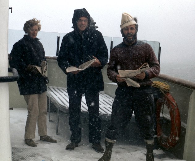 Three people standing on the deck of a boat with snow falling around them.