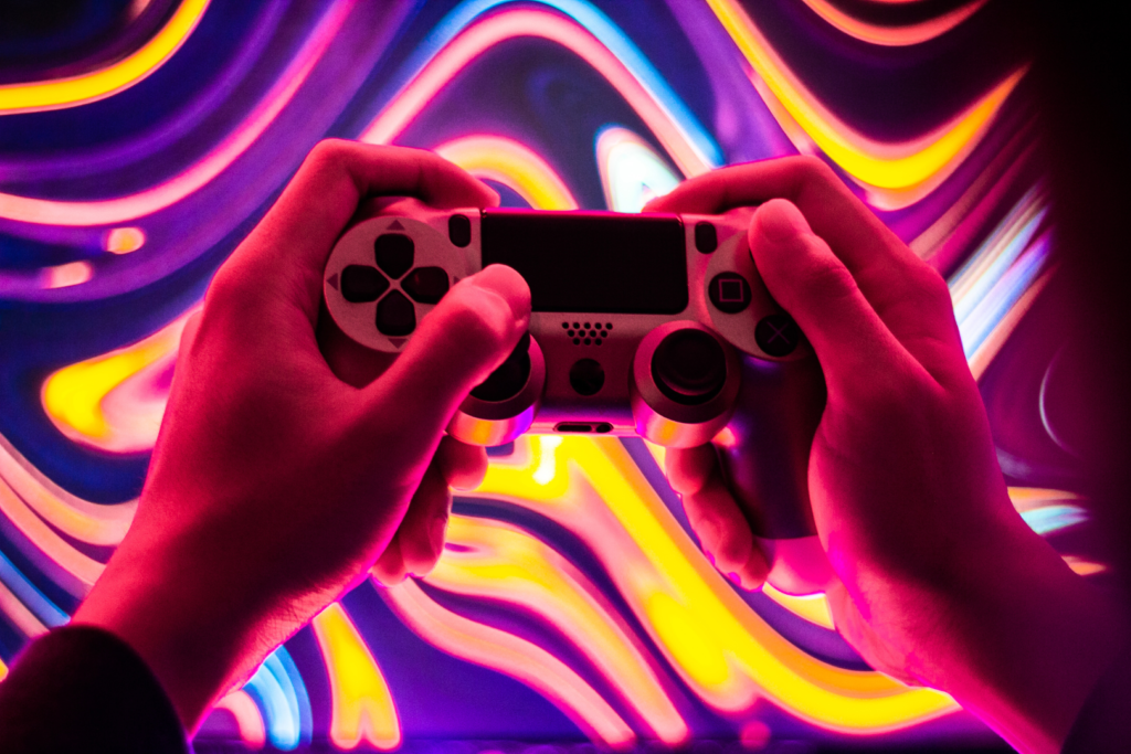 A person's hands holding a playstation video game controller. Behind the controller the background is covered in neon curved lights.