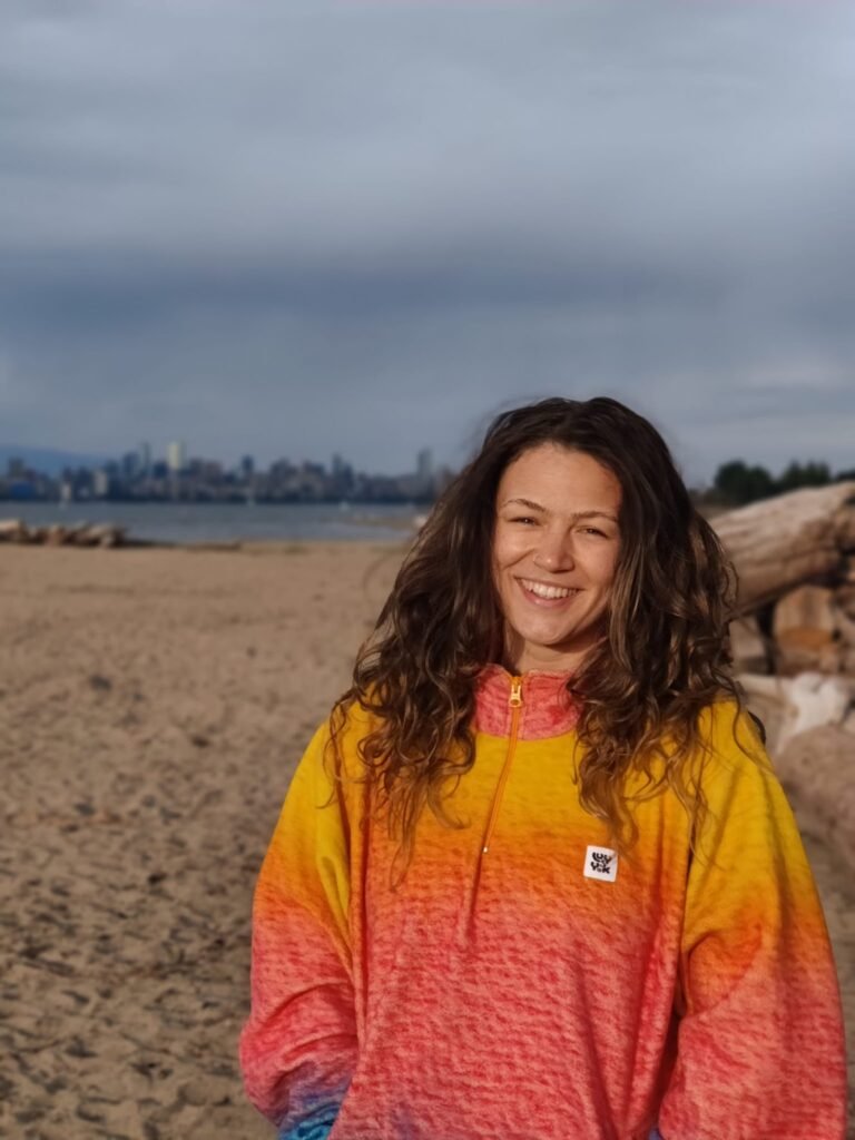 Woman in a yellow and orange jacket standing on a beach smiles at the camera