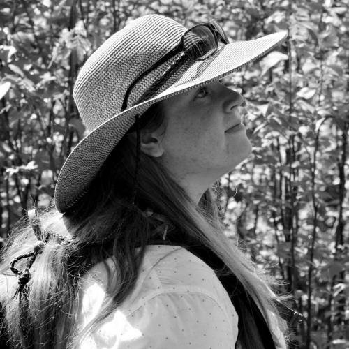 A person with long hair and a floppy hat looks into the distance. Photo is black and white.